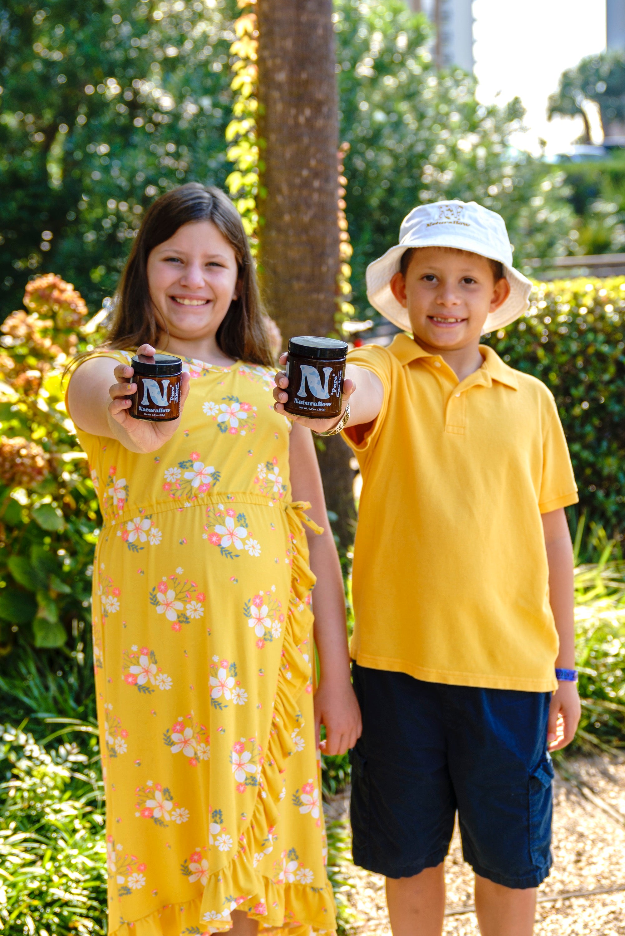 A picture of two young children standing next to each other and smiling while extending their arm and holding Naturallow's Pure line of products. On the left is a young girl and on the right is a young boy