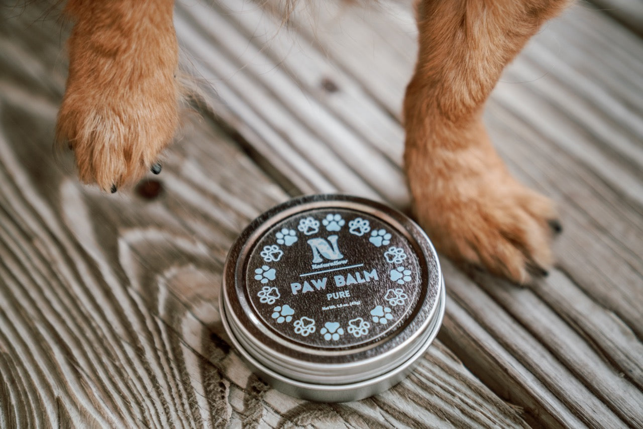A photo of a small, circular tin sitting on woodgrain in between a set of brown dog paws. The tin has purple paw print illustrations. Its label reads "Naturallow Paw Balm Lavender. Net Weight 1.5 oz (42g)"
