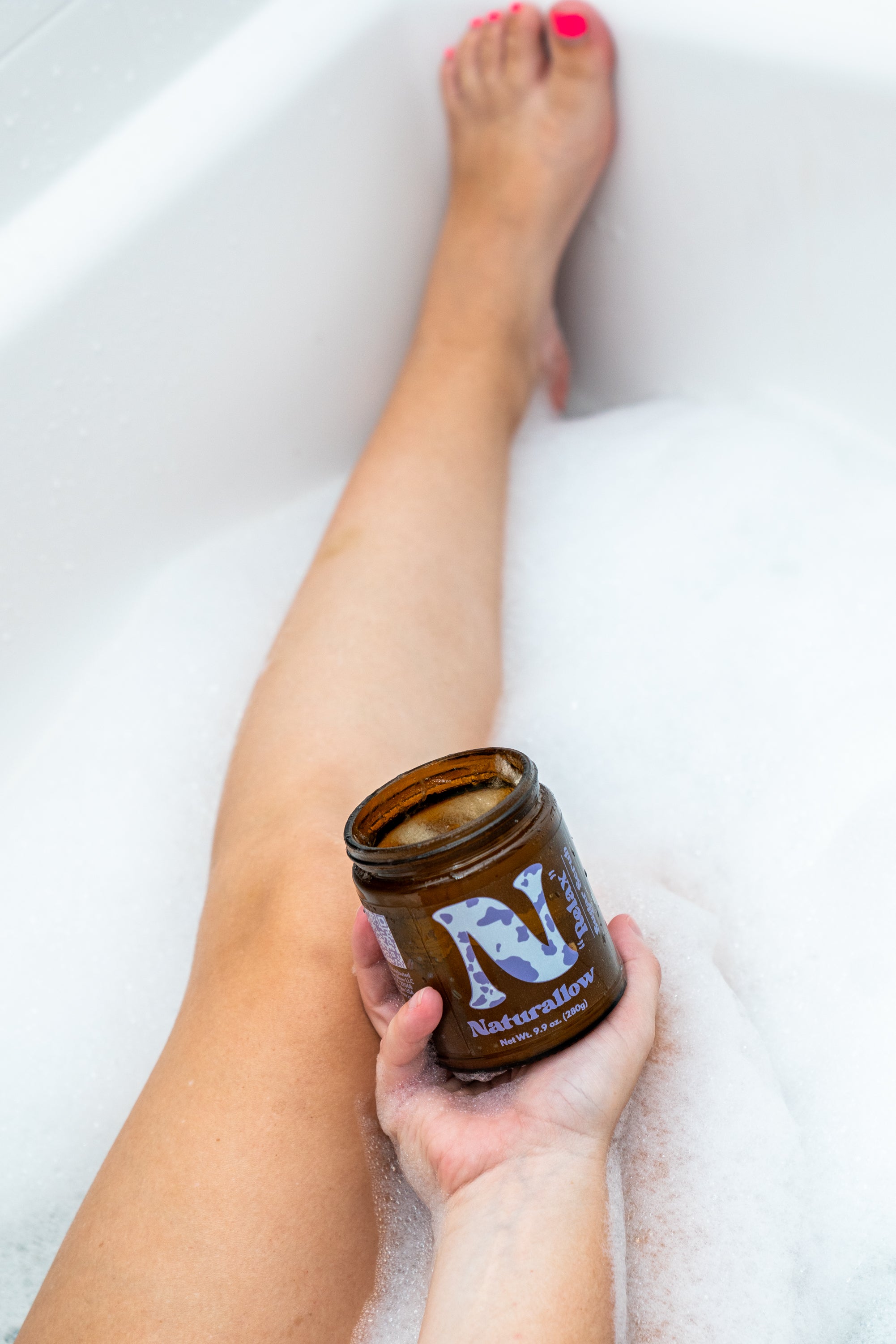 A Photo showing a leg in a bubble bath with a hand holding out Naturallow's Relax Sugar Scrub